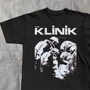 May include: Black t-shirt featuring the band name "THE KLINIK" in white capital letters above a grayscale graphic of figures. The text "...est treasures" is below the band name. A music-themed item.