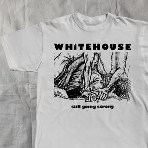 以下が含まれることがあります： 白の半袖Tシャツ。黒いグラフィックとテキストが特徴です。グラフィックは人物のスケッチで、テキストは画像の上に「WHITEHOUSE」、下に「still going strong」と書かれています。