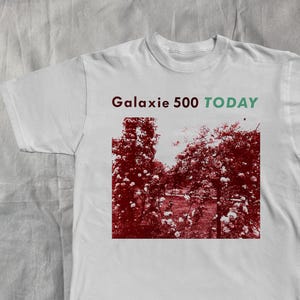 Könnte beinhalten: Weißes T-Shirt mit einem rot-weißen Blumengrafik. Der Text "Galaxie 500 TODAY" ist in Grün und Rot über der Grafik aufgedruckt. Das Shirt besteht aus einem weichen Material.