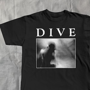 Op de afbeelding: Zwart t-shirt met het woord "DIVE" in witte blokletters boven een zwart-wit afbeelding van een vage figuur. Het shirt is gemaakt van een zacht materiaal.
