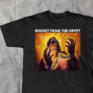 May include: Black t-shirt featuring a vibrant graphic of a screaming ape-like figure with chains, set against a bright yellow and orange background. The text "ROCKET FROM THE CRYPT" is printed in white above the graphic.