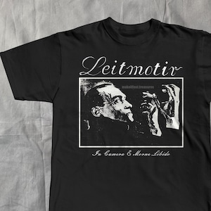 May include: Black t-shirt with a black and white graphic of a man, the word "Leitmotiv" above the image, and the text "In Camera & Morne Libido" below. The shirt is made of a soft material.