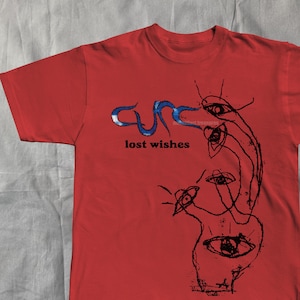May include: Red t-shirt featuring the band name "Cure" in blue and white, with the words "lost wishes" in black. The shirt has a black graphic design of eyes and abstract shapes.