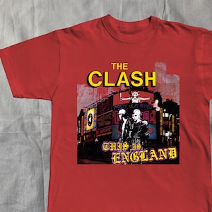 Może przedstawiać: Czerwona koszulka z grafiką zespołu The Clash. Projekt zawiera nazwę zespołu w kolorze żółtym oraz napis "THIS IS ENGLAND". Grafika przedstawia również członków zespołu i krajobraz miejski.