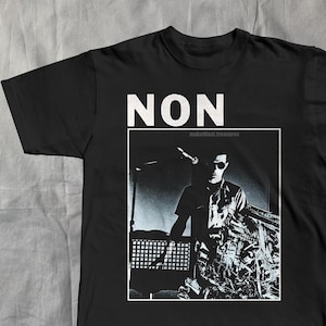 May include: Black t-shirt with the word "NON" in white block letters at the top. Below the text is a black and white image of a person wearing sunglasses. The text "makefast.treasures" is above the image.