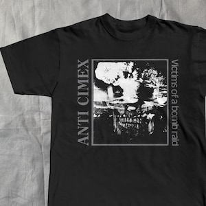 Op de afbeelding: Zwart t-shirt met een vervaagde afbeelding van een schedel en de tekst "ANTI CIMEX" verticaal aan de linkerkant en "Victims of a bomb raid" aan de rechterkant. Het ontwerp is in tinten wit en grijs.