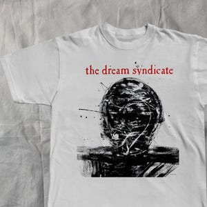 May include: White t-shirt featuring the red text "the dream syndicate" above a black and white abstract graphic. The graphic appears to be a stylized face or head with splatters.