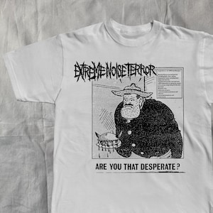May include: White t-shirt featuring a black and white graphic. The graphic includes the text "EXTREME NOISE TERROR" above an illustration of a man and the text "ARE YOU THAT DESPERATE?"