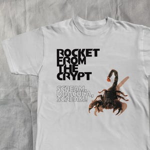 Könnte beinhalten: Weißes T-Shirt mit dem Text "ROCKET FROM THE CRYPT" und einer Skorpion-Grafik. Darunter steht der Satz "SCREAM, DRACULA, SCREAM!". Der Skorpion ist schwarz mit einem roten Stachel.