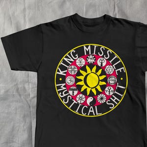 May include: Black t-shirt with a circular design. The text "KING MISSILE SHIT" encircles a yellow sun, with various symbols in red, white, and black. A bold graphic tee.