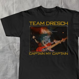 May include: Black t-shirt featuring a band image with the text "TEAM DRESCH" above and "CAPTAIN MY CAPTAIN" below. The image shows a person playing a red guitar. The text and image are in a gold color.