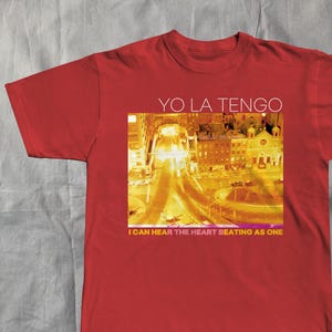 May include: Red t-shirt with the text "YO LA TENGO" above a yellow and orange city scene graphic. The text "I CAN HEAR THE HEART BEATING AS ONE" is below the image. A casual, everyday garment.