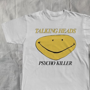May include: White t-shirt featuring the text "TALKING HEADS" above a yellow smiley face with the words "PSYCHO KILLER" below. The design is printed in black and yellow on a white, short-sleeved shirt.