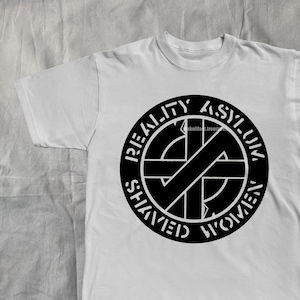 Op de afbeelding: Wit T-shirt met een zwarte afbeelding. De afbeelding is een cirkel met de woorden "REALITY ASYLUM SHAVED WOMEN" rond een zwart-wit kruisontwerp. Het T-shirt ligt plat op een lichtgrijze ondergrond.