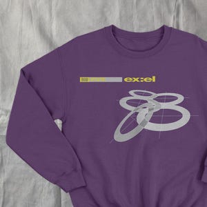May include: A purple sweatshirt with long sleeves. The design features the word "ex:el" in yellow and a series of overlapping silver rings. The sweatshirt is made of a soft material.