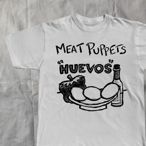 May include: White t-shirt with a black graphic design. The design includes the text "Meat Puppets" above the word "Huevos", and a plate with eggs, a pepper, and a bottle. The design is in a hand-drawn style.