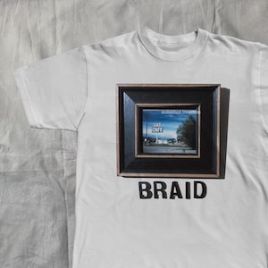 以下が含まれることがあります： ガソリンスタンドの額入り画像と「BRAID」の文字が黒いブロック体でプリントされた白いTシャツ。額入り画像はTシャツの中央に配置されています。Tシャツは明るい灰色の表面に平らに置かれています。