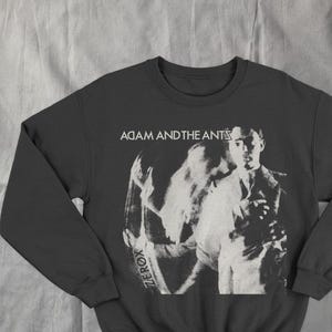 Può includere: Felpa grigio scuro con un'immagine in bianco e nero di un uomo e il testo "ADAM AND THE ANTS" e "ZEROX". La felpa ha maniche lunghe e girocollo.