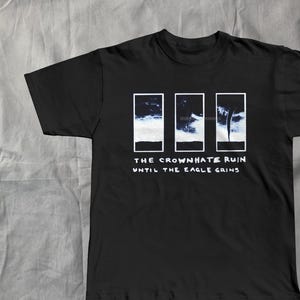 Könnte beinhalten: Schwarzes T-Shirt mit weißem Grafikdesign. Das Design zeigt drei rechteckige Tafeln mit einem Bild eines bewölkten Himmels und dem Text "THE CROWNHATE RUIN" und "UNTIL THE EAGLE GAINS" in weißen Blockbuchstaben.