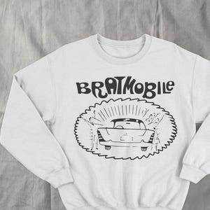 May include: White sweatshirt with the text "BRATMOBILE" above a black and white illustration of a vintage car. The car is surrounded by a sunburst design. The sweatshirt has long sleeves and a crew neck.