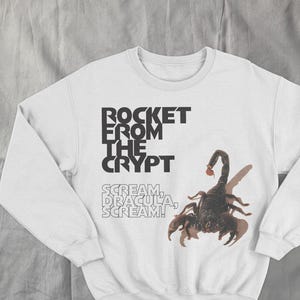 May include: White sweatshirt featuring a black scorpion graphic and the text "ROCKET FROM THE CRYPT" above "SCREAM, DRACULA, SCREAM!". The design is printed on a long-sleeved, crew neck sweatshirt.
