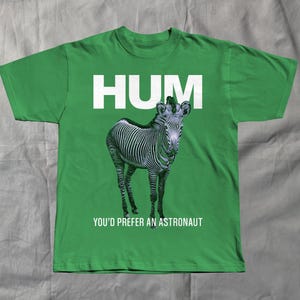May include: Green t-shirt with the word "HUM" in white block letters above a zebra illustration. Below the zebra, the text reads "YOU'D PREFER AN ASTRONAUT". The shirt is made of a soft material.