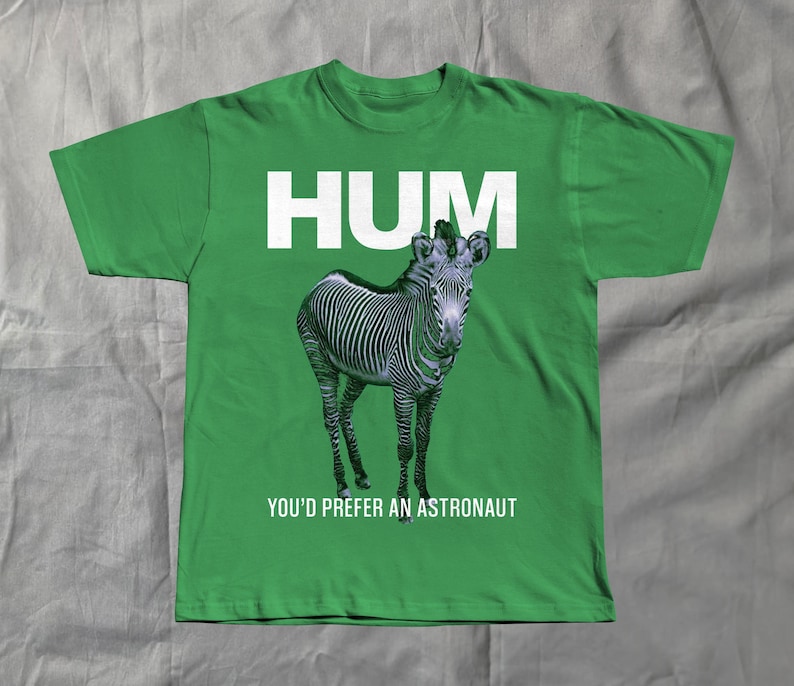 May include: Green t-shirt with the word "HUM" in white block letters above a zebra illustration. Below the zebra, the text reads "YOU'D PREFER AN ASTRONAUT". The shirt is made of a soft material.