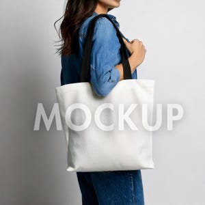 May include: A white tote bag with black straps is held by a person wearing a denim shirt and jeans. The word "MOCKUP" is printed in large, light gray letters across the front of the bag. The bag is made of a sturdy material.