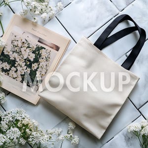 May include: A cream-colored tote bag with black handles, placed on a white tiled surface. A magazine with a floral image and the word "MOCKUP" is next to the bag. White flowers are scattered around the items.