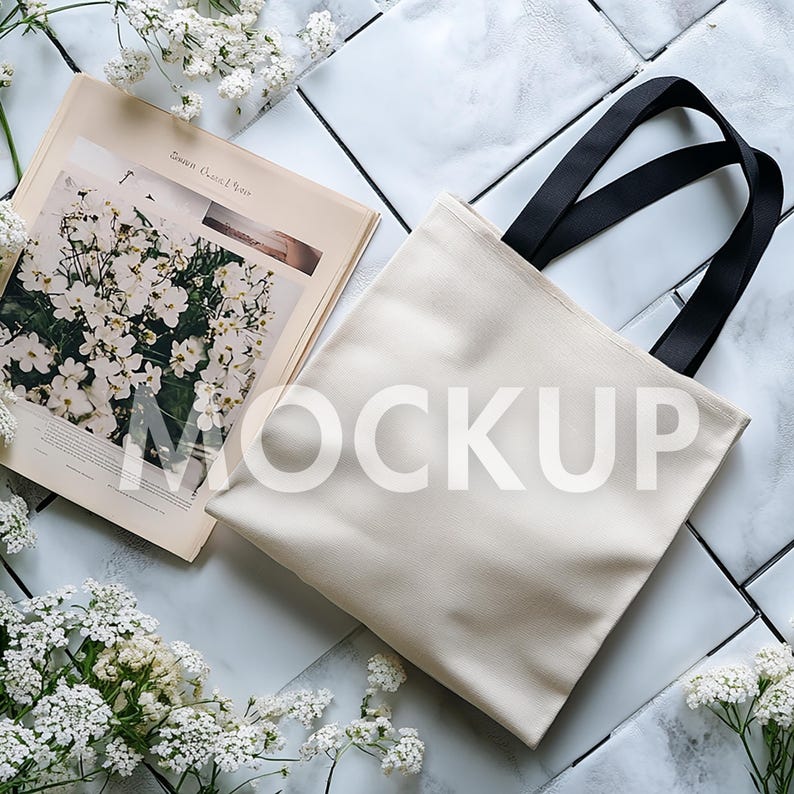May include: A cream-colored tote bag with black handles, placed on a white tiled surface. A magazine with a floral image and the word "MOCKUP" is next to the bag. White flowers are scattered around the items.