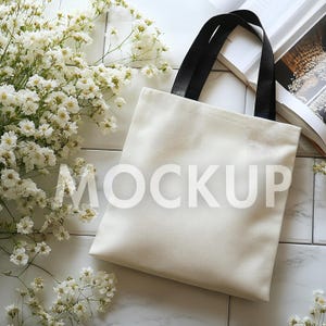 May include: A cream-colored tote bag with black handles, resting on a white tiled surface. The word "MOCKUP" is printed on the front of the bag. White flowers and a partially visible open book are also in the image.