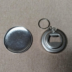 May include: A silver-colored bottle opener with a key ring and a matching round metal disc. The bottle opener has a circular design with a bottle cap opener. The disc is a solid, round metal piece.