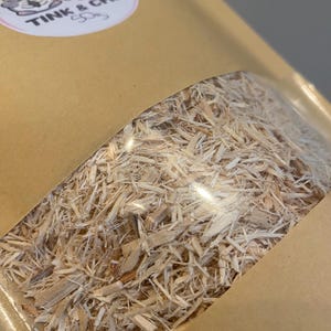 May include: A brown paper bag with a clear window showing a large amount of light brown wood shavings inside. The bag has a sticker on the front that says "Tink & Chip".