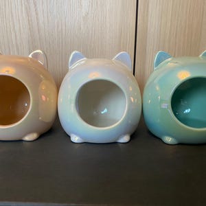 May include: Three ceramic cat-shaped pet beds in different colours: light brown, light blue and light green. Each bed has a round opening for the pet to enter.