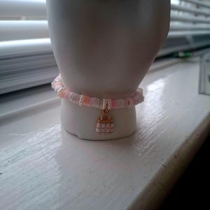 May include: A pink and white beaded bracelet with a gold-colored purse charm. The bracelet is made of small, flat, round beads in shades of pink and white, with small white beads interspersed. The charm is a miniature purse.