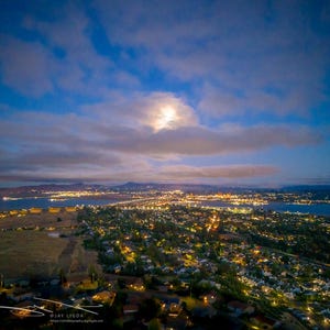 May include: An aerial view of a city at dusk, with a full moon peeking through the clouds. The city lights are illuminated, reflecting on the water. The sky is a mix of blue and orange hues.
