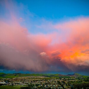 May include: A vibrant landscape photograph featuring a dramatic sky with pink and orange clouds against a bright blue sky. The foreground shows a town nestled between green hills.