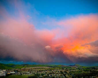 May include: A vibrant landscape photograph featuring a dramatic sky with pink and orange clouds against a bright blue sky. The foreground shows a town nestled between green hills.