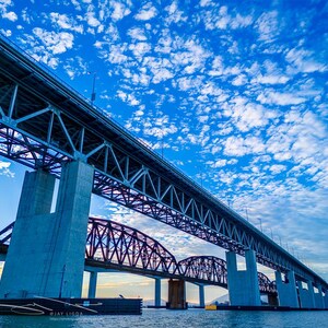 May include: A low-angle shot of a large bridge with a steel truss design, supported by concrete pillars. The sky is a vibrant blue with scattered white clouds. A second bridge with a dark arch design is visible in the background.
