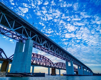 May include: A low-angle shot of a large bridge with a steel truss design, supported by concrete pillars. The sky is a vibrant blue with scattered white clouds. A second bridge with a dark arch design is visible in the background.