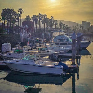 The rising sun glows above boats moored in a marina, casting a silver reflection across the calm waters.