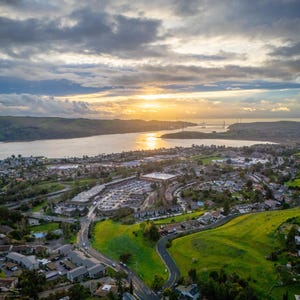 On a partly cloudy evening, the sun sets above the Carquinez Strait, illuminating the town and the green hills below.