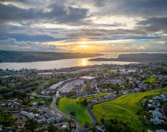 May include: An aerial view of a suburban landscape featuring residential buildings, winding roads, and green hillsides leading to a wide body of water. The horizon shows a distant bridge under a cloudy sky with a bright golden sunset glow.