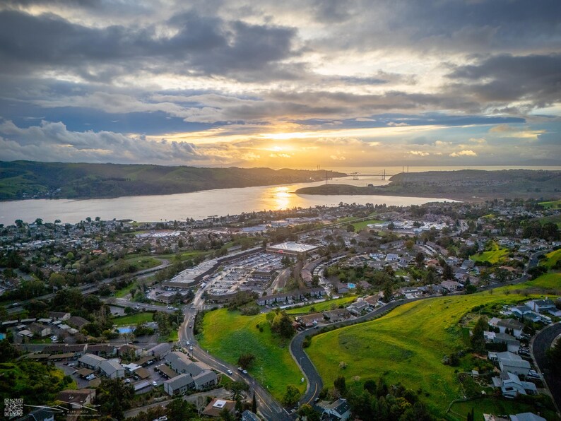 On a partly cloudy evening, the sun sets above the Carquinez Strait, illuminating the town and the green hills below.