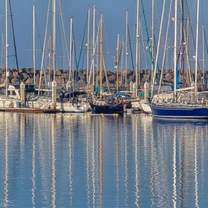 The masts of sailing ships reflect in the calm waters while the boats remain docked beneath a clear blue sky.