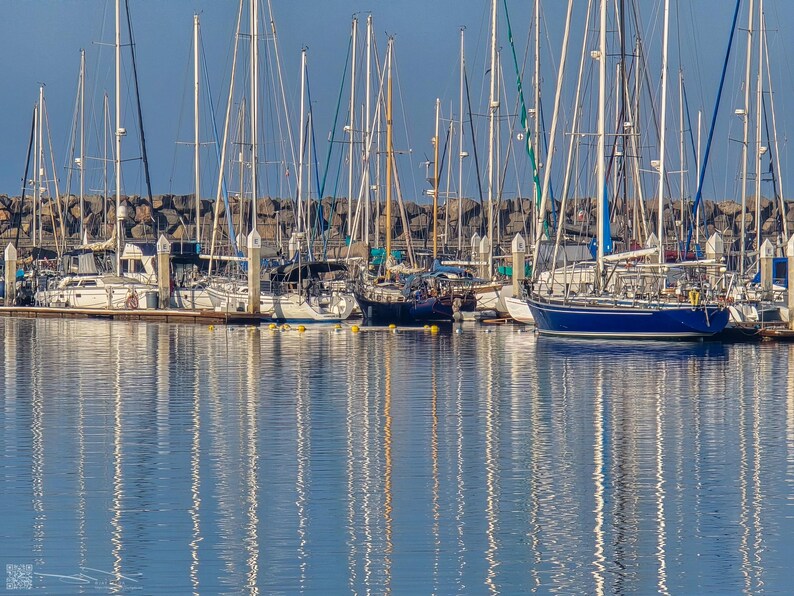 The masts of sailing ships reflect in the calm waters while the boats remain docked beneath a clear blue sky.