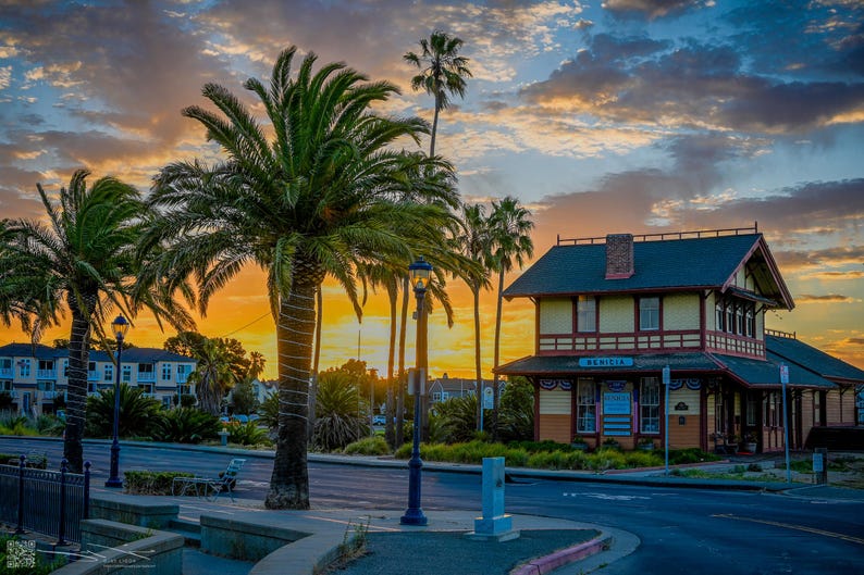 The sun rises behind the Depot Building in Benicia, CA, casting a warm yellow glow across the horizon. The palm trees stand their ground, ready to greet the new day.