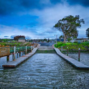 May include: A concrete boat ramp leads into the water, flanked by two long, narrow wooden docks. The left dock features a wooden railing, while the right dock is open. The scene is set under a cloudy, deep blue sky with green grassy banks.