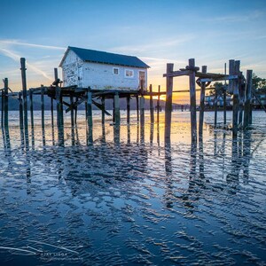 May include: A scenic landscape featuring a weathered, white building on stilts over water at sunset. Wooden pilings support the structure, reflecting in the rippled water. The sky transitions from blue to orange, creating a tranquil coastal scene.