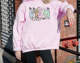 May include: Pink hoodie with a graphic design that says "He is Risen" with a cross, flowers, and bunny ears. The text "Matthew 28:6" is also included.